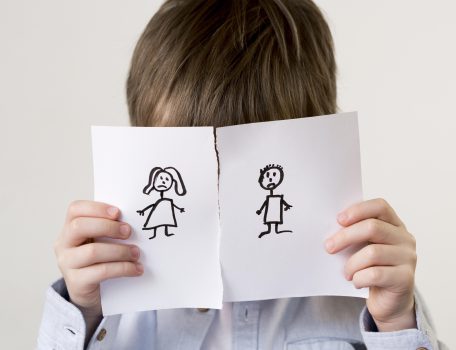 Image shows a young boy with dark hair and a blue shirt with his face covered by a piece of paper torn in half with a stick drawing of a woman and man on each half for the Family Justice Council Blog