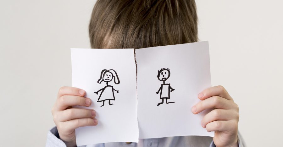 Image shows a young boy with dark hair and a blue shirt with his face covered by a piece of paper torn in half with a stick drawing of a woman and man on each half for the Family Justice Council Blog